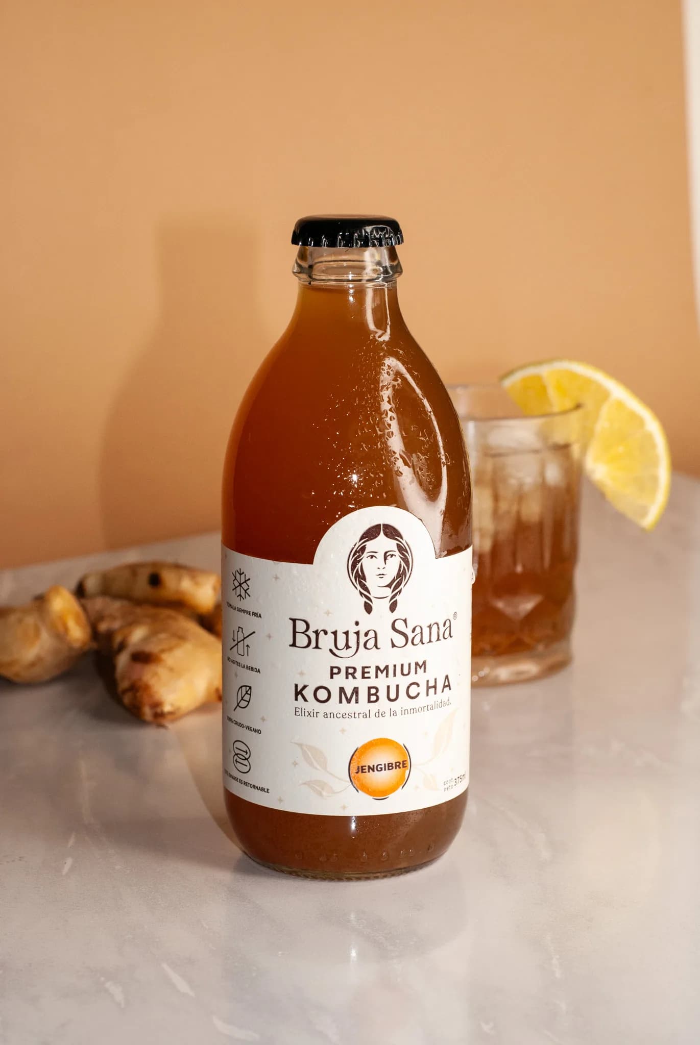 Botella de Kombucha Bruja Sana sabor Jengibre junto a un vaso con hielo y limón y raíz de jengibre